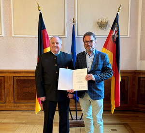 Zwei Personen mit Urkunde vor Flaggen