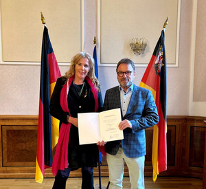 Zwei Personen mit Urkunde vor Flaggen