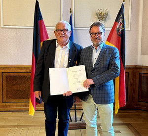 Zwei Personen mit Urkunde vor Flaggen
