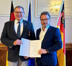 Zwei Männer mit Urkunde vor Flaggen