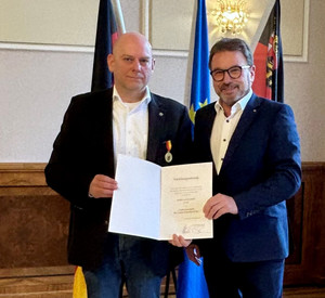 Zwei Männer mit Urkunde vor Flaggen