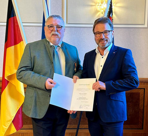 Zwei Männer mit Urkunde vor Flaggen